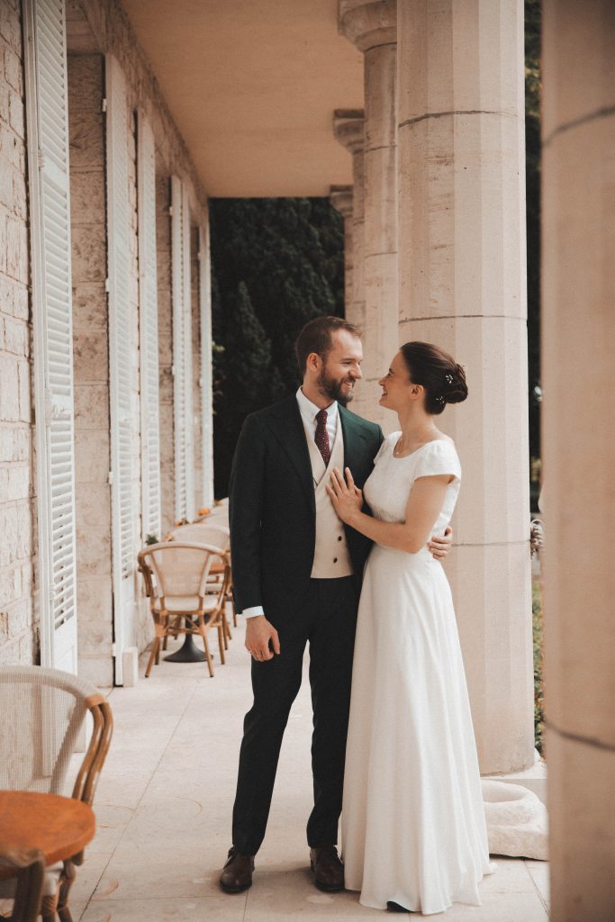 Couple de mariés élégants devant un mur blanc aux volets, robe sobre et costume bordeaux, photo mariage
